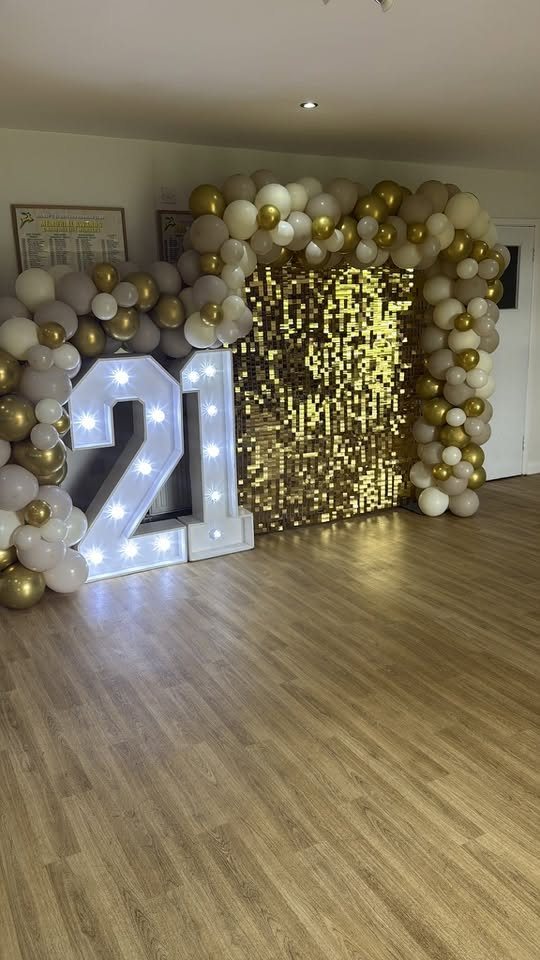 A 21st birthday set-up with light-up numbers, gold sequin wall and balloon arch