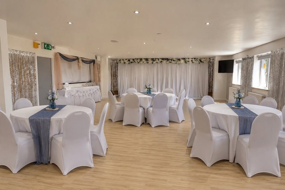 The hall laid out with round tables, white chair covers and a floral backdrop for a wedding
